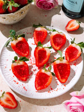 Champagne strawberry jello shots arranged on a white plate with fresh strawberries, pink roses, and a prosecco bottle.