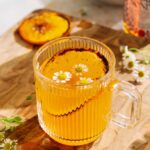 Brûléed orange hot toddy with amaro in a clear glass mug, garnished with chamomile flowers and a caramelized orange slice on a wooden board.