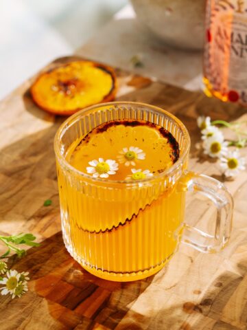 Brûléed orange hot toddy with amaro in a clear glass mug, garnished with chamomile flowers and a caramelized orange slice on a wooden board.