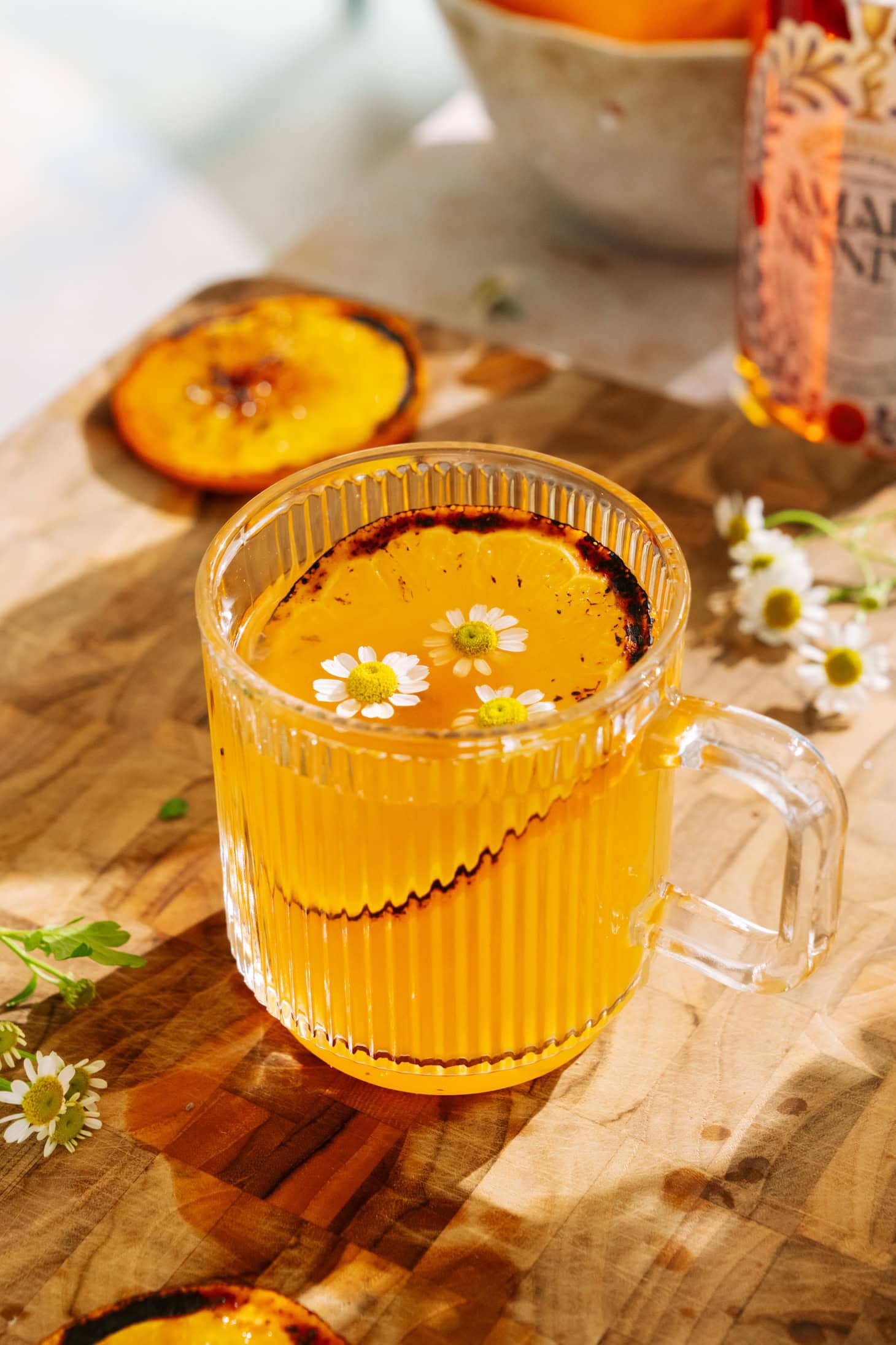 Brûléed orange hot toddy with amaro in a clear glass mug, garnished with chamomile flowers and a caramelized orange slice on a wooden board.