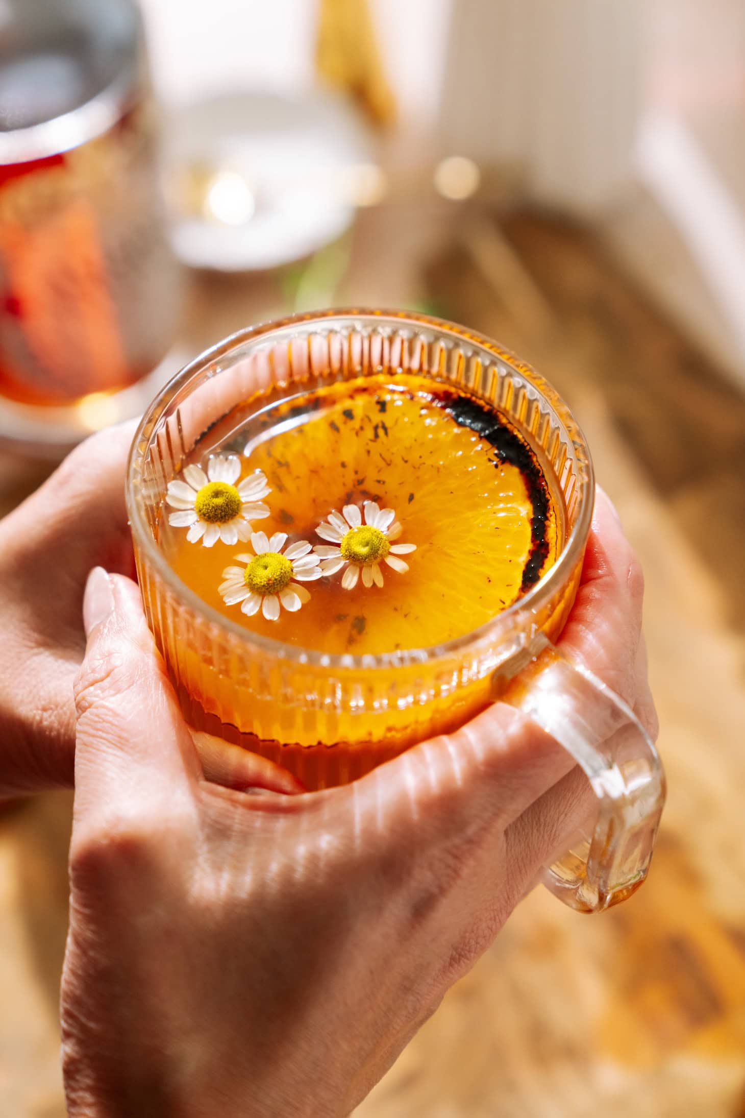 Hands holding a warm glass mug of brûléed orange hot toddy with amaro, topped with chamomile flowers and a charred citrus slice.