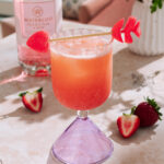 Blushy strawberry gin cocktail with foam and heart candy garnish, styled with roses, daisies, and fresh strawberries on a sunlit table.