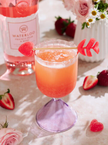 Strawberry Valentine’s cocktail in a purple stemmed glass with ice, garnished with heart-shaped candy skewers, fresh strawberries, and a pink gin bottle in the background.