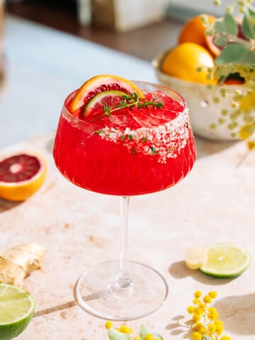 Bright red ginger blood orange mocktail in a coupe glass with a salted rim, garnished with citrus slices and fresh thyme on a sunlit countertop.