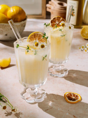Two creamy limoncello spritz drinks garnished with dried lemon, herbs, and flowers on a light surface
