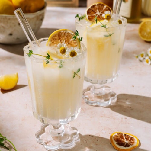 Two creamy limoncello spritz drinks garnished with dried lemon, herbs, and flowers on a light surface