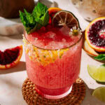 Bright coconut blood orange margarita with crushed ice, fresh mint, lime wheel, and citrus sugar rim in a textured glass, surrounded by sliced blood oranges and limes.