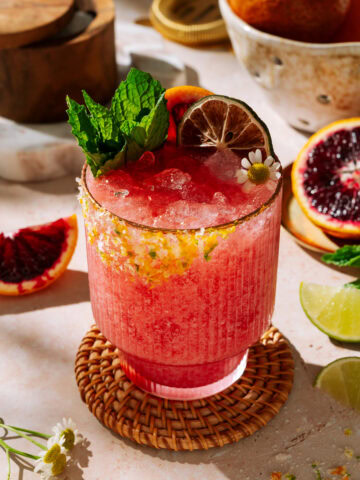 Bright coconut blood orange margarita with crushed ice, fresh mint, lime wheel, and citrus sugar rim in a textured glass, surrounded by sliced blood oranges and limes.