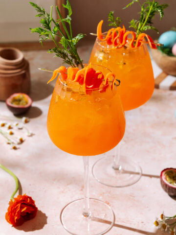 Passion Fruit Spritz cocktail with carrot garnish and pastel Easter eggs on spring table.
