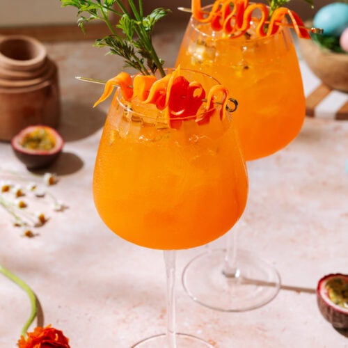 Passion Fruit Spritz cocktail with carrot garnish and pastel Easter eggs on spring table.