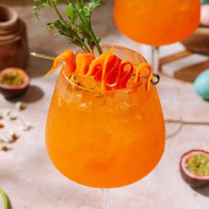 Passion Fruit Spritz cocktail with carrot ribbon garnish and fresh herbs in wine glasses.