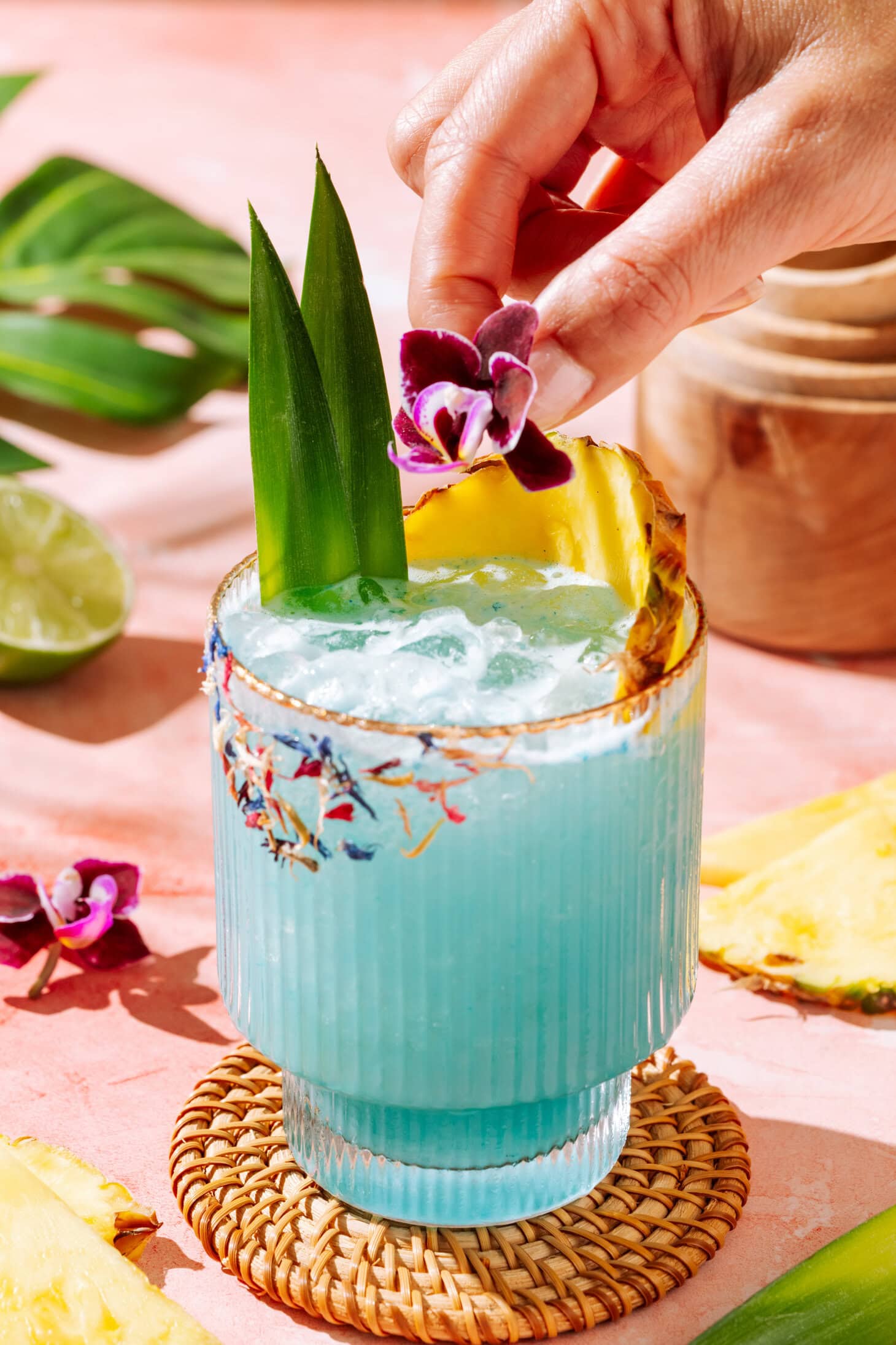 Blue tropical cocktail with pineapple garnish, crushed ice, and floral rim on a pink background with a hand placing an edible flower on the cocktail as a decoration.