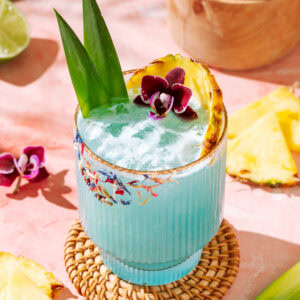 Blue tropical cocktail with pineapple garnish, crushed ice, and floral rim on a pink background