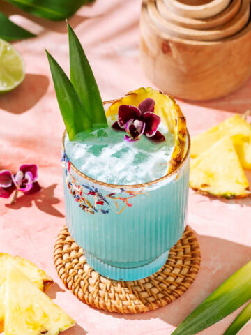 Blue tropical cocktail with pineapple garnish, crushed ice, and floral rim on a pink background
