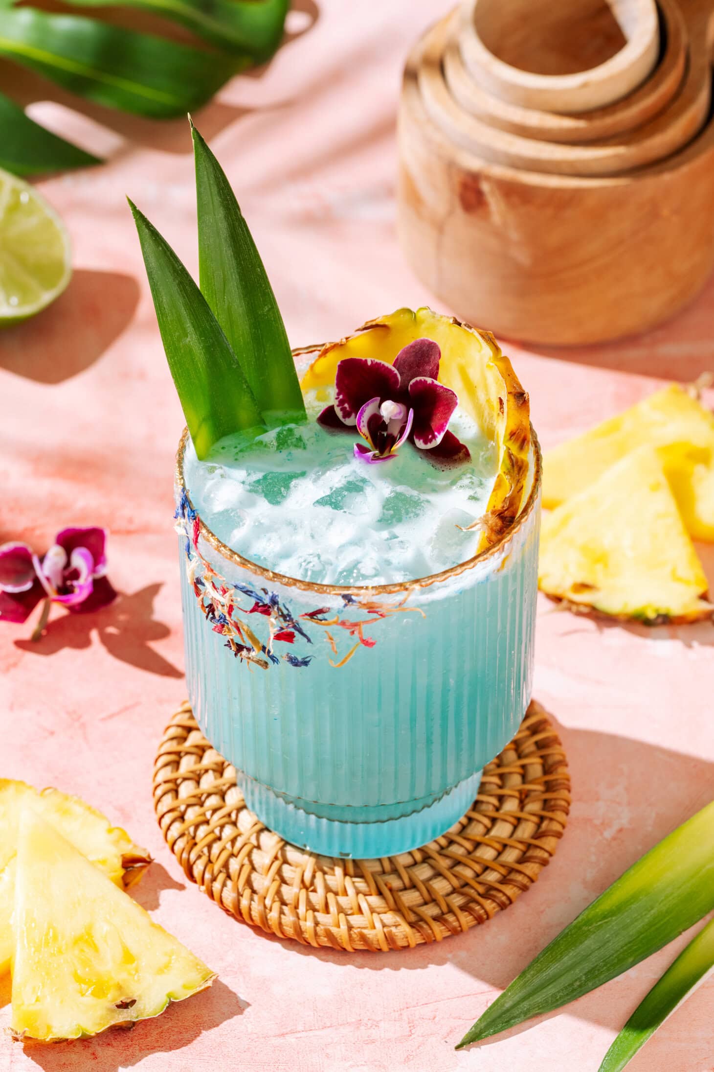 Blue tropical cocktail with pineapple garnish, crushed ice, and floral rim on a pink background