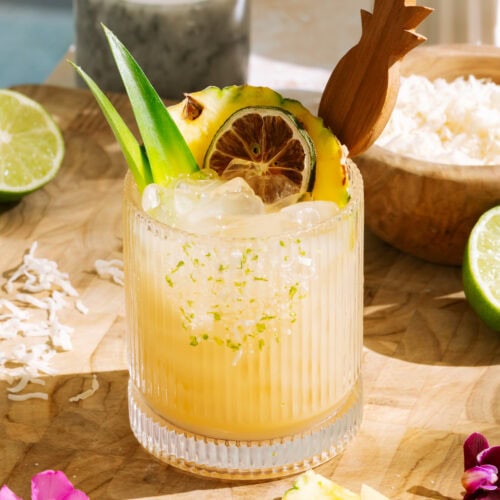 Tropical pineapple coconut mocktail with crushed ice, lime zest rim, dried citrus wheel, and fresh pineapple garnish, served in a textured glass with limes and coconut flakes.