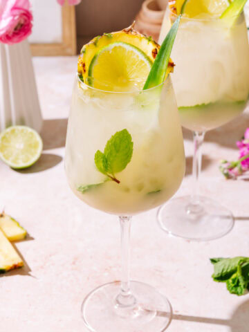 Piña Colada Hugo Spritz served in a tall wine glass with pineapple, lime, fresh mint, and tropical garnish served over ice.