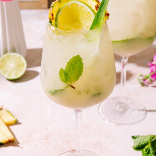 Piña Colada Hugo Spritz served in a tall wine glass with pineapple, lime, fresh mint, and tropical garnish served over ice.