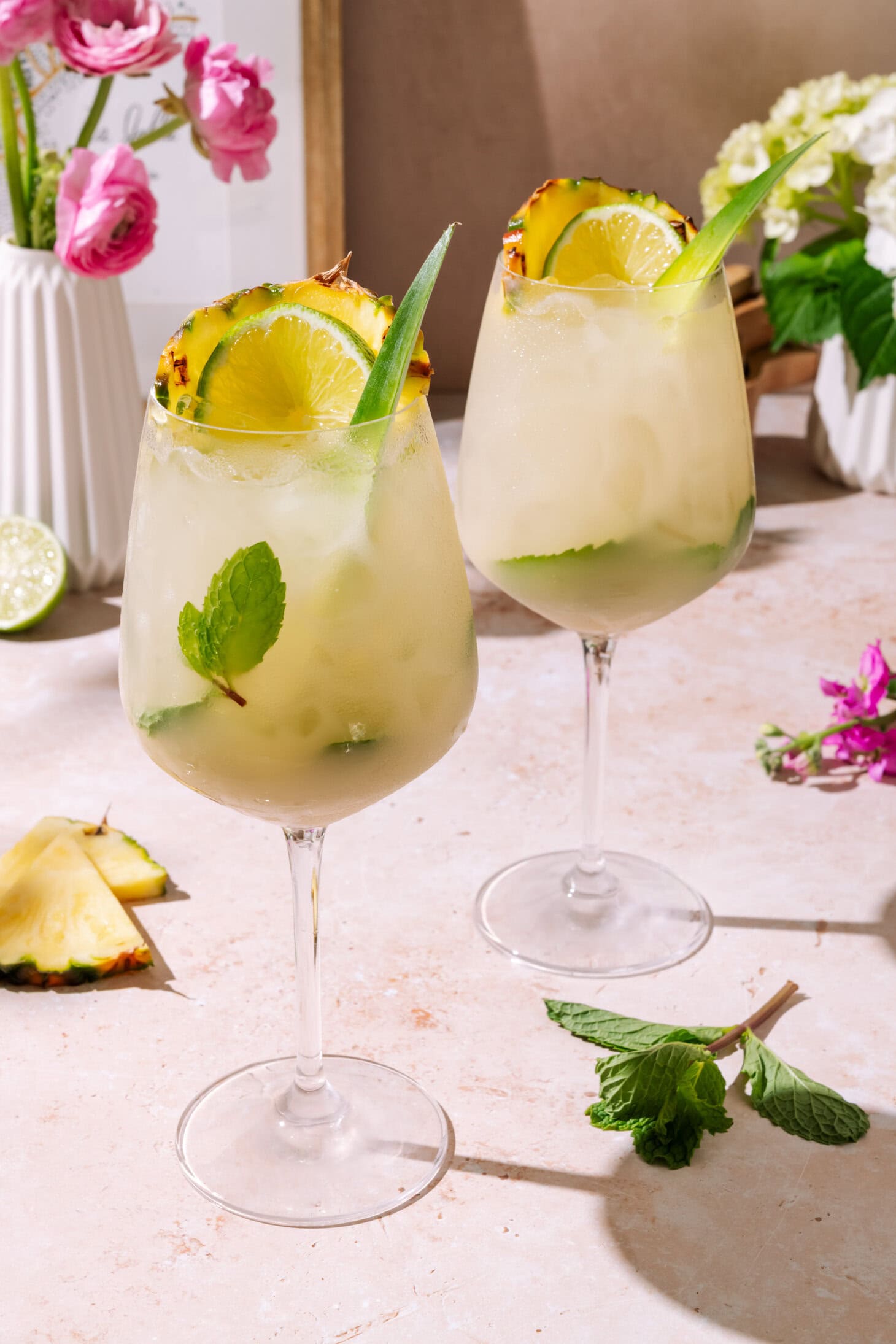 Piña Colada Hugo Spritz served in 2 tall wine glasses with pineapple, lime, fresh mint, and tropical garnish served over ice.