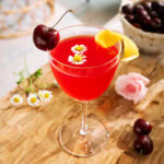 Cherry Bee’s Knees mocktail garnished with fresh cherries, lemon peel, and delicate chamomile flowers.