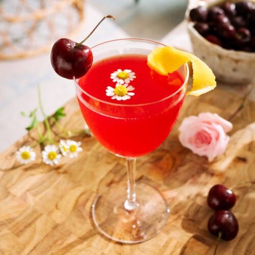 Cherry Bee’s Knees mocktail garnished with fresh cherries, lemon peel, and delicate chamomile flowers.