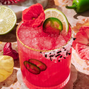 Vibrant spicy hibiscus margarita with lime, jalapeño slices, and edible flower garnish, served over crushed ice in a salt-rimmed glass.