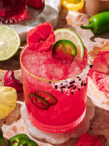 Vibrant spicy hibiscus margarita with lime, jalapeño slices, and edible flower garnish, served over crushed ice in a salt-rimmed glass.