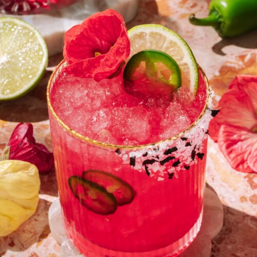Vibrant spicy hibiscus margarita with lime, jalapeño slices, and edible flower garnish, served over crushed ice in a salt-rimmed glass.