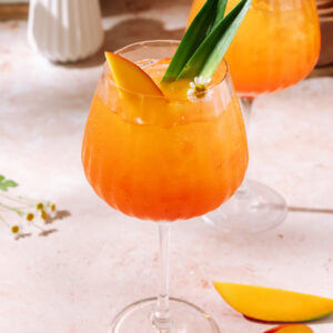 Aperol Mango Spritz with fresh mango slices, tropical leaf garnish, and edible flowers over ice.