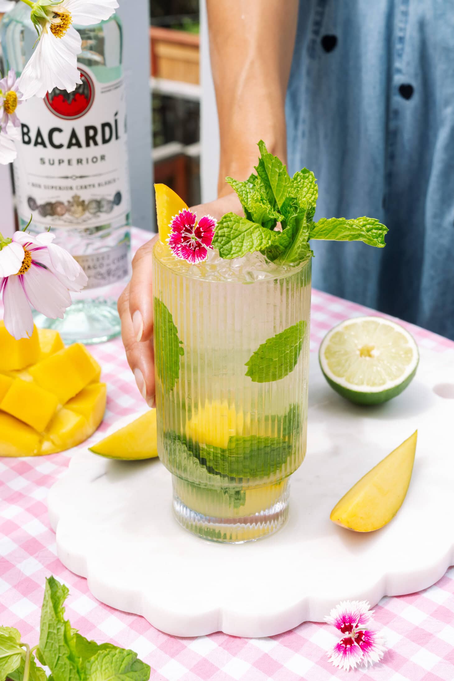Mango Mojito in a tall ribbed glass, garnished with fresh mango, mint, lime, and pink flowers.