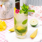 Mango Mojito in a tall ribbed glass, garnished with fresh mango, mint, lime, and pink flowers.