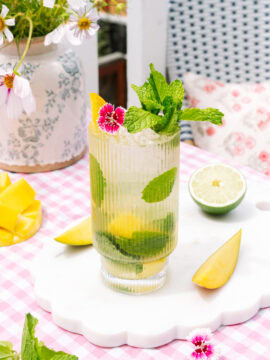 Mango Mojito in a tall ribbed glass, garnished with fresh mango, mint, lime, and pink flowers.