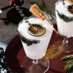 Frozen Coconut Margarita in a stemmed glass with a black salt rim, dried lime, lavender garnish and small broom as a Halloween decoration.