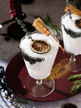 Frozen Coconut Margarita in a stemmed glass with a black salt rim, dried lime, lavender garnish and small broom as a Halloween decoration.