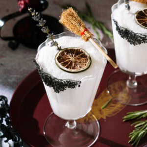 Frozen Coconut Margarita in a stemmed glass with a black salt rim, dried lime, lavender garnish and small broom as a Halloween decoration.