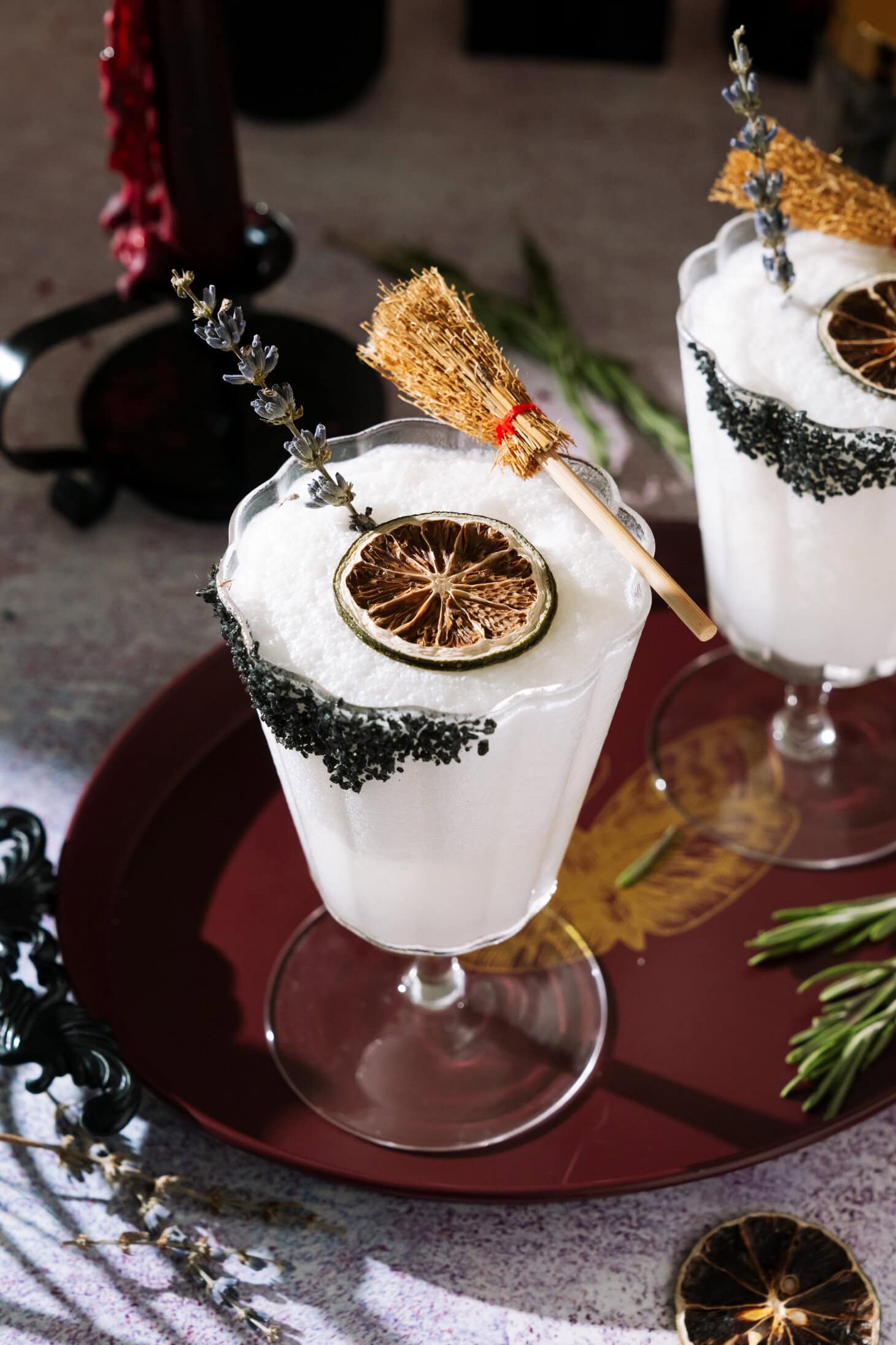 Frozen Coconut Margarita in a stemmed glass with a black salt rim, dried lime, lavender garnish and small broom as a Halloween decoration.