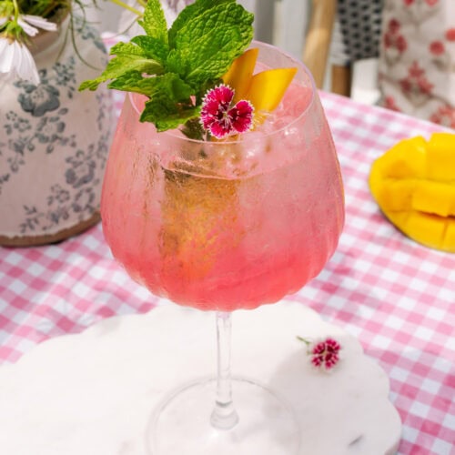Sarti Spritz in tall glass garnished with fresh mango, mint, and pink flowers.