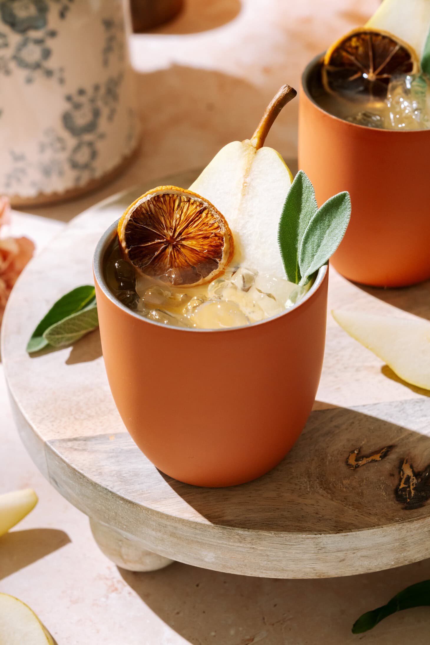 A close-up of a Amaretto Pear Margarita in a terracotta cup, garnished with pear slices and dried citrus on a wooden board.