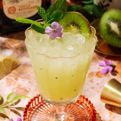 Tequila Kiwi Cocktail in a stemmed glass, garnished with fresh kiwi, mint, and a purple flower.