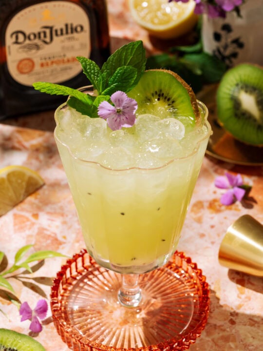Tequila Kiwi Cocktail in a stemmed glass, garnished with fresh kiwi, mint, and a purple flower.