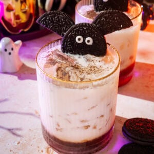 Two Halloween-themed drinks garnished with Oreo cookies shaped like bats, surrounded by Halloween decorations and cookies on a festive table.