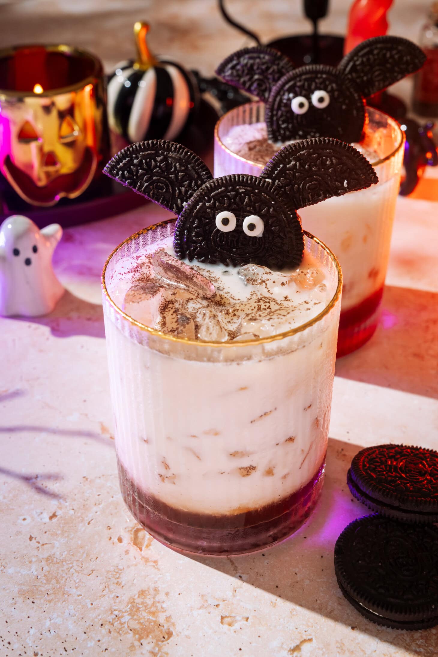 Two Halloween-themed drinks garnished with Oreo cookies shaped like bats, surrounded by Halloween decorations and cookies on a festive table.