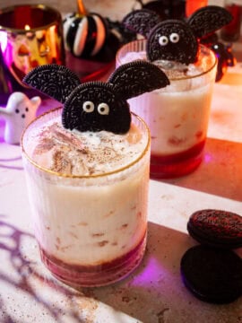 Two Halloween-themed drinks garnished with Oreo cookies shaped like bats, surrounded by Halloween decorations and cookies on a festive table.
