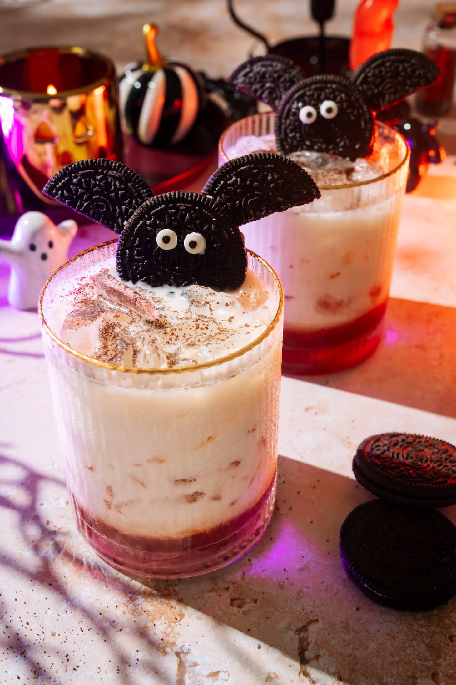 Two Halloween-themed drinks garnished with Oreo cookies shaped like bats, surrounded by Halloween decorations and cookies on a festive table.