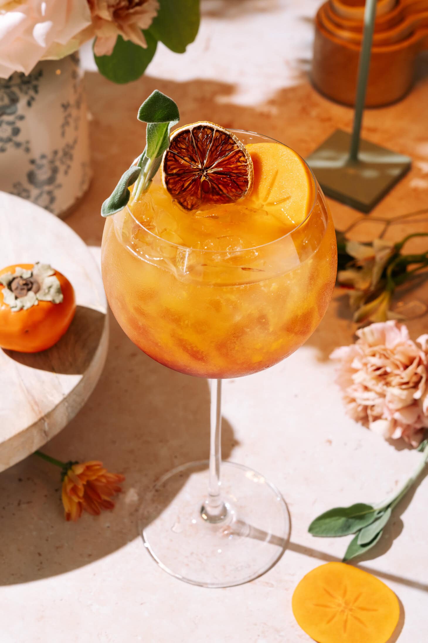 Easy Persimmon Pomegranate Mocktail served in a stemmed glass with dried citrus garnish, surrounded by flowers and decor on a sunny table.
