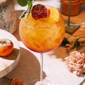 Easy Persimmon Pomegranate Mocktail served in a stemmed glass with dried citrus garnish, surrounded by flowers and decor on a sunny table.